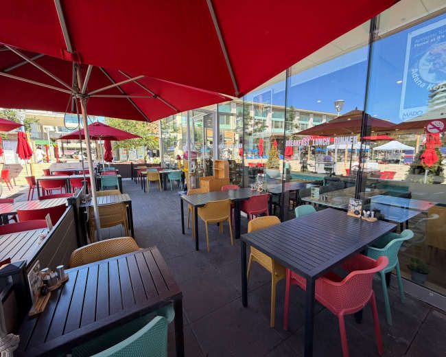 The scene shows an outdoor patio of a restaurant with colorful tables and chairs, shaded by large red umbrellas.