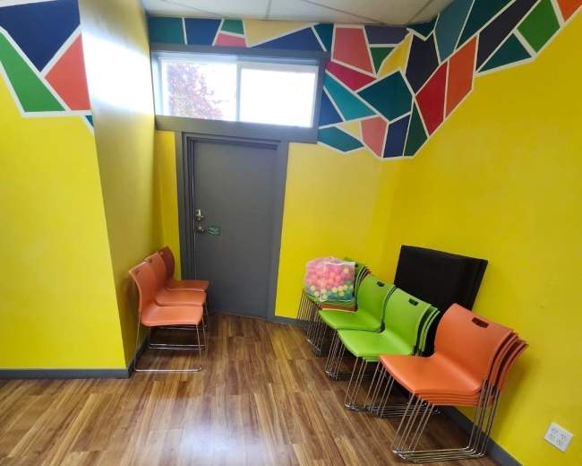 The image shows a brightly colored room with yellow walls, featuring a row of colorful chairs and a stack of blankets near a closed door.