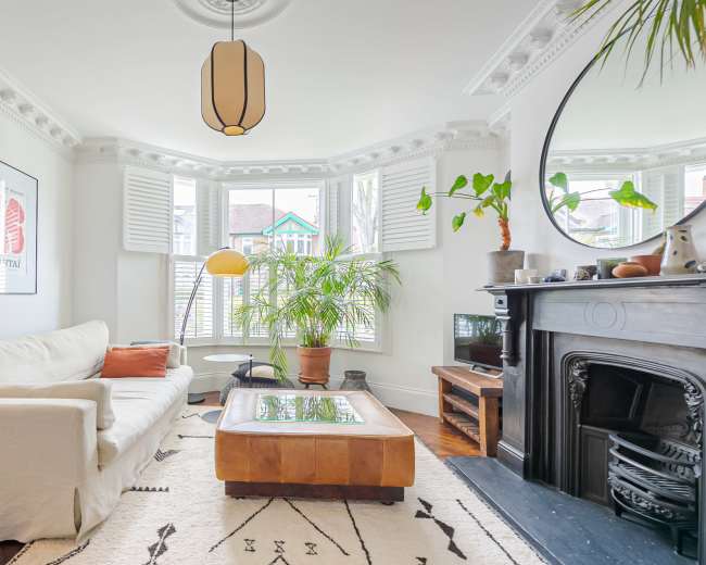 A living room features a white sofa, a large coffee table, and green plants, with bay windows and decorative molding.