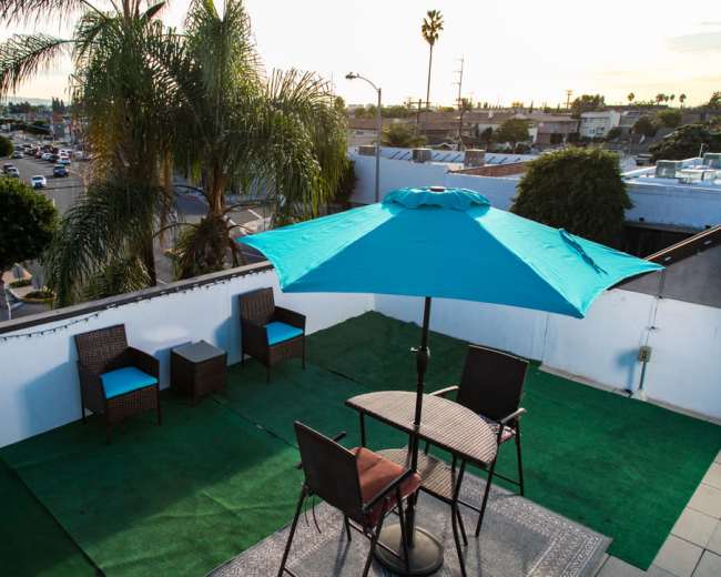 A rooftop area features a turquoise umbrella over a table and chairs, surrounded by palm trees and a view of city streets.