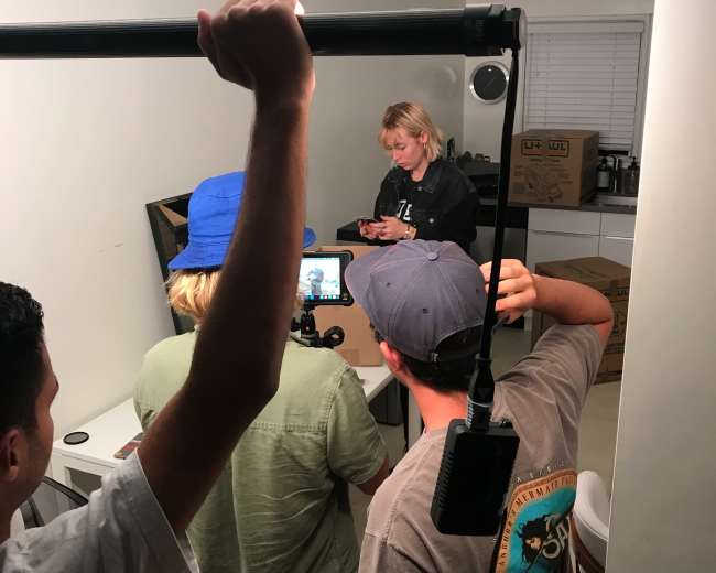 A group of four people is filming in a small room, focused on a laptop screen while one person holds a camera stabilizer.