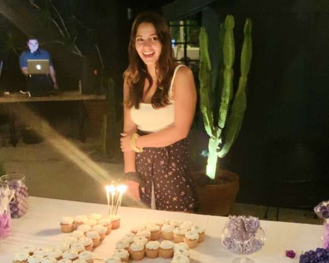 A woman stands beside a table decorated with cupcakes and a "30" made of cupcakes, with candles lit on top, in a dimly lit outdoor setting.