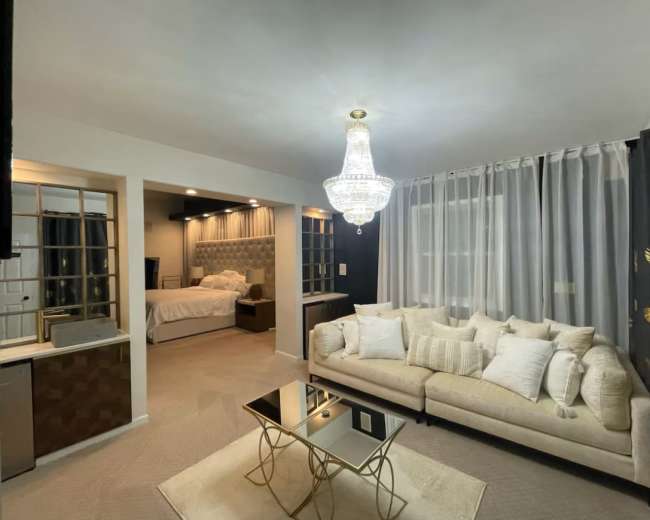 The image shows a well-lit room featuring a modern sofa with decorative pillows, a glass coffee table, and a view of a bedroom area in the background.