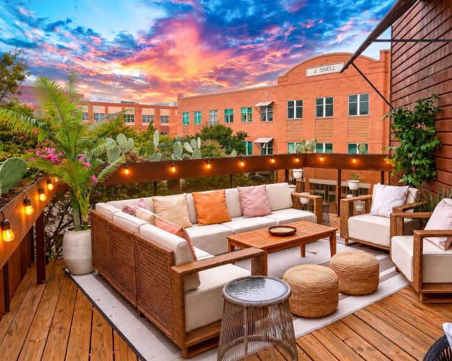 A modern patio features comfortable seating, a wooden table, and decorative plants against a vibrant sunset sky.