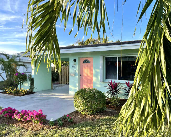 The image shows a light green house with a pink door, surrounded by tropical plants and flowers, under a clear blue sky.