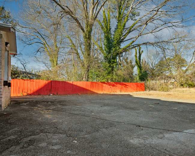 The image shows an empty paved area bordered by a red wooden fence and surrounded by tall trees.