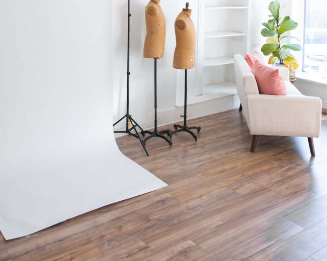 The image shows a bright, minimalistic studio space featuring two dressmaker's mannequins, a white backdrop on a stand, and a light-colored sofa with a cushion.