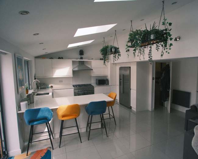 The image shows a modern kitchen with a central island, three bar stools in various colors, stainless steel appliances, and hanging plants.