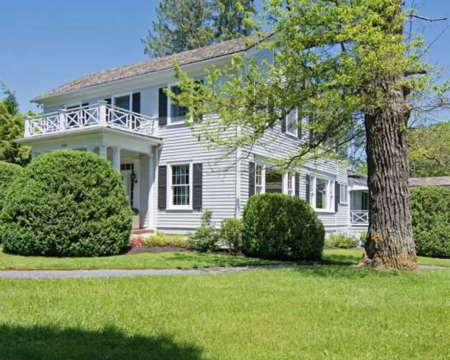 A two-story white house with a porch and neatly trimmed shrubs is surrounded by green grass and trees.
