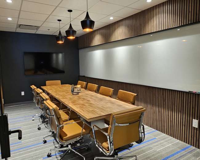 The image shows a modern conference room with a long wooden table, orange leather chairs, a blank whiteboard, and a large screen on the wall.
