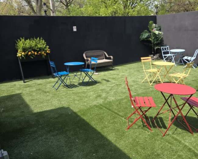 The image shows a landscaped outdoor area with artificial grass and various colored metal chairs and tables, surrounded by greenery and a black wall.