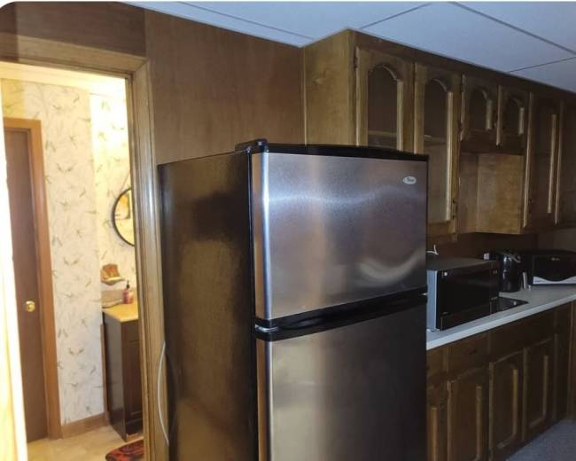 The image shows a kitchen area with a stainless steel refrigerator, wooden cabinetry, a countertop with appliances, and a doorway leading to another room.