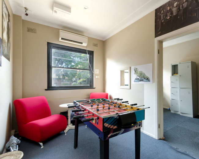 The image shows a small, tidy room with a foosball table, a red couch, and an open doorway leading to another room featuring filing cabinets.