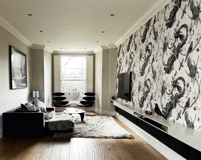 The image shows a modern living room with a large window, a black sofa, two black lounge chairs, a television mounted on a wall, and a textured wall featuring a floral pattern.