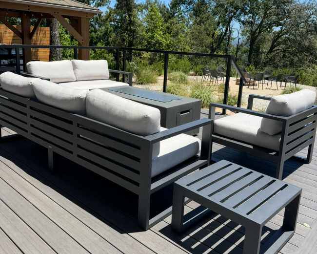 The image shows a modern outdoor seating area with gray furniture and a small side table on a wooden deck surrounded by trees.