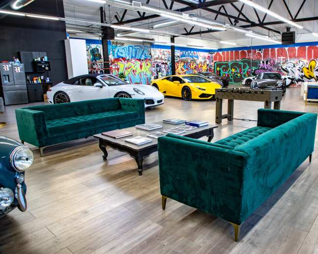 The image shows a spacious garage with modern cars, green velvet sofas, a coffee table with magazines, and vibrant graffiti on the walls.