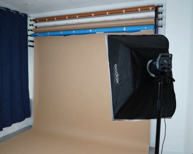 The image shows a photography studio setup with a brown backdrop, professional lighting equipment, and rolled backdrops on the wall.