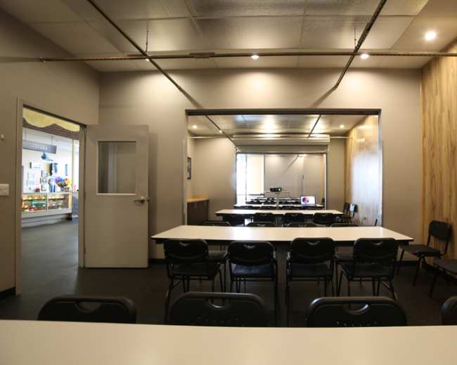 A spacious meeting room featuring several black chairs arranged around white tables, with a large projection screen visible in the background.