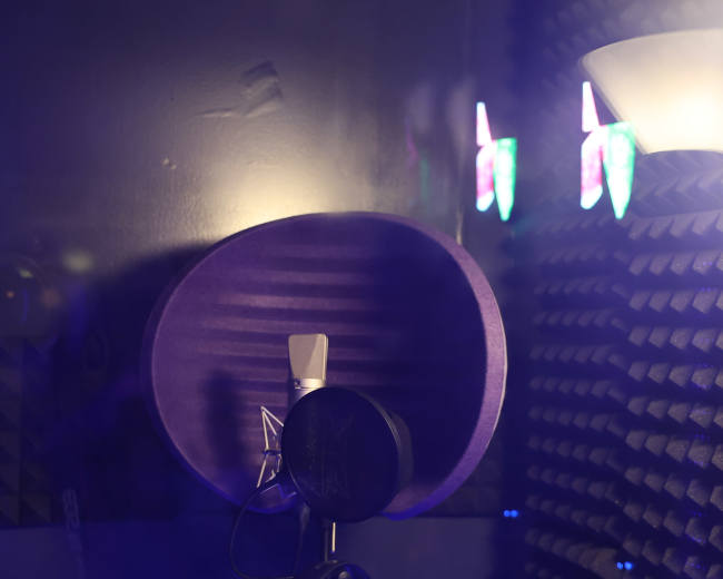A microphone is positioned in a soundproof recording booth with acoustic panels and colorful lights in the background.