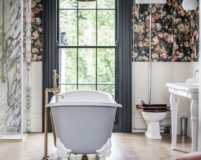 The image shows a bathroom with a clawfoot bathtub, a shower area with marble walls, and floral wallpaper alongside a window providing natural light.