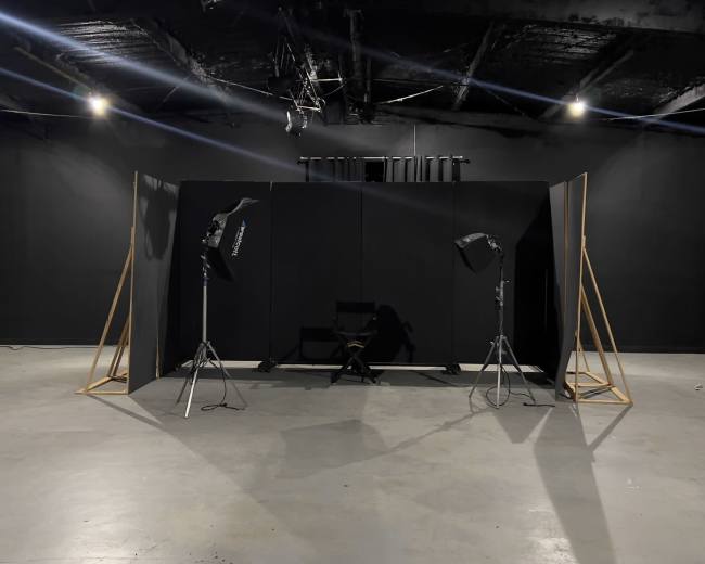 A minimalist photography studio setup with black walls, a director's chair, and lighting equipment positioned around a central backdrop.