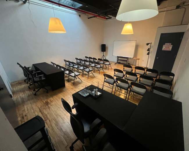 The image shows a spacious, empty room set up for a meeting, featuring a long table at the front, several rows of chairs, and a whiteboard on one side.