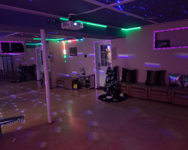 A spacious room with colorful lights, a projector on the ceiling, a Christmas tree in one corner, and a seating area with metallic pillows.