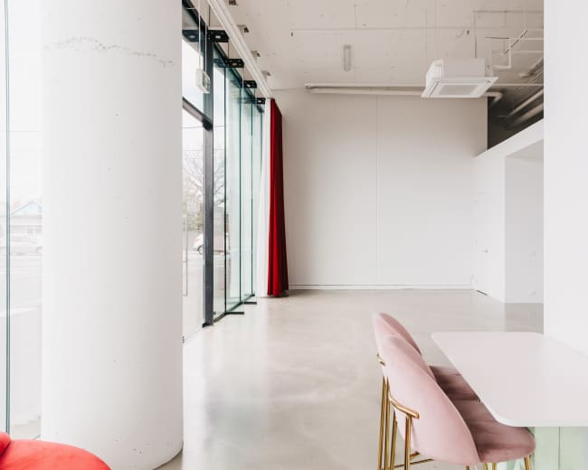 The image shows a modern, minimalist interior featuring a pink chair and a small table near large glass windows with red curtains.