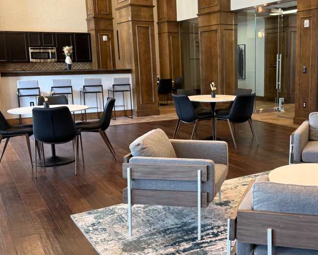 The image depicts a stylish communal area featuring various seating arrangements, including gray chairs and tables, with a foosball table in the corner and a large window allowing natural light.