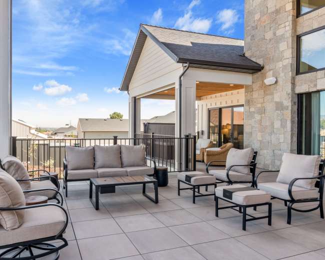 The image shows a spacious outdoor patio furnished with several seating options, including a sofa, armchairs, and small tables, surrounded by a railing and with a blue sky overhead.