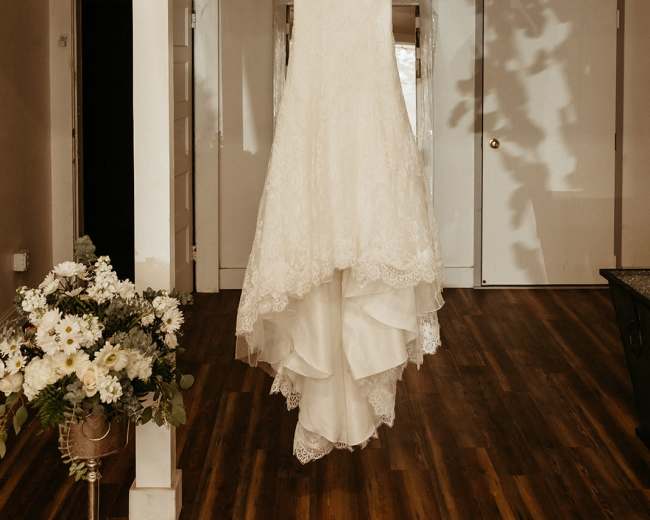 A wedding dress is displayed on a hanger in a softly lit room with a flower arrangement on a table.