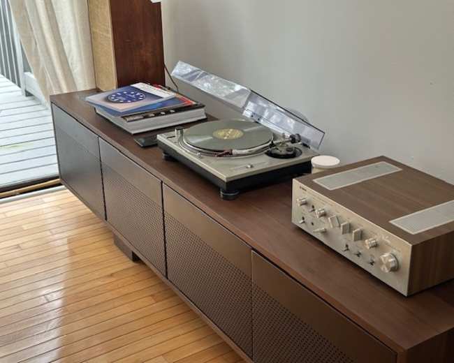 The image features a wooden console with a vinyl record player and an audio receiver, accompanied by several records on top.