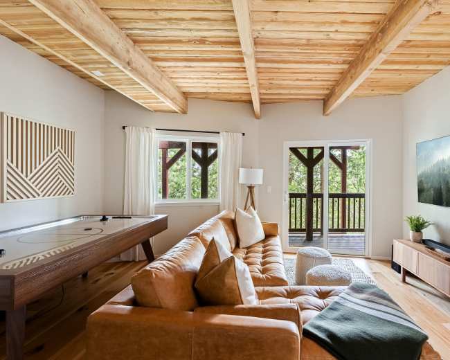 The living room features a large leather sectional couch, a wooden table, and a wall-mounted television, with large windows providing a view of the outdoors.