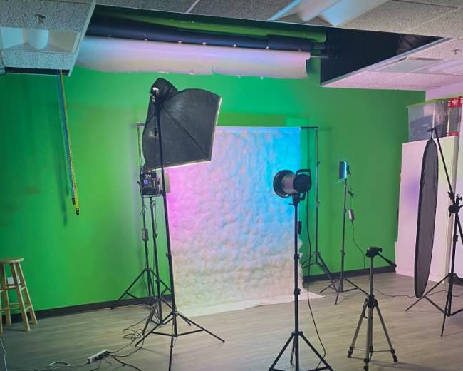 A photography studio with a green wall, equipped with softbox lights, a backdrop stand, and various photography equipment arranged on the floor.