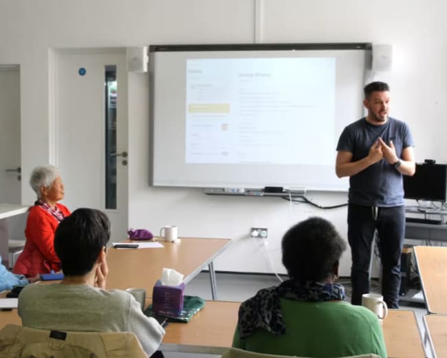 A man stands and speaks in front of a group of seated elderly individuals during a presentation in a bright room.