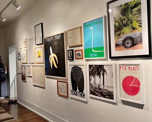 The image shows a gallery wall adorned with a variety of framed artworks, including prints, photographs, and posters in different sizes and styles.