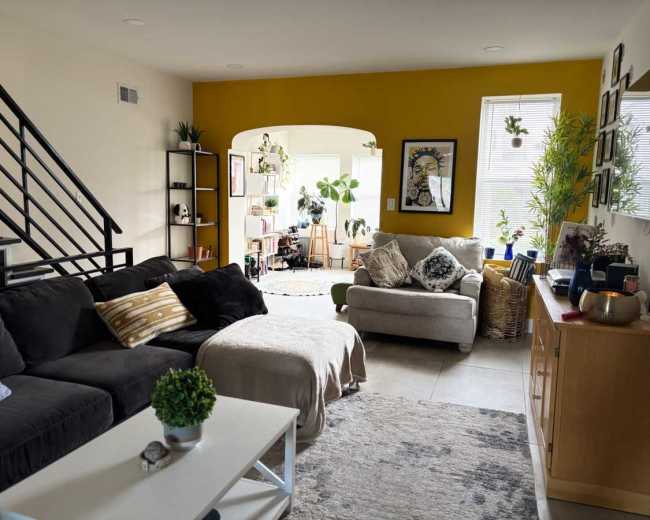 The image shows a bright and open living area featuring a dark sofa, a light-colored armchair, and a wooden media console, with plants and artwork adorning the walls.