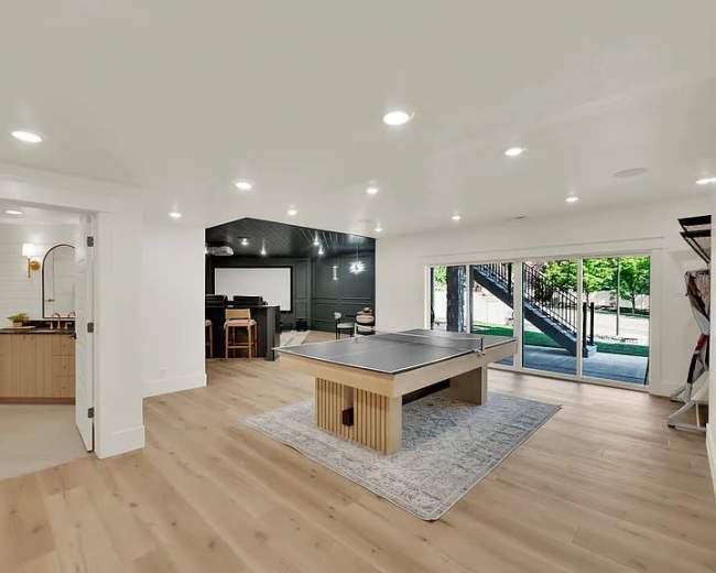 The image shows a modern basement interior featuring a pool table on a rug, with sliding glass doors leading outside, and a kitchenette area visible to the left.