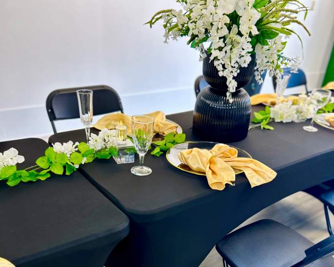 A black tablecloth covers a table set with glassware and yellow napkins, complemented by a floral centerpiece and greenery along the edge.
