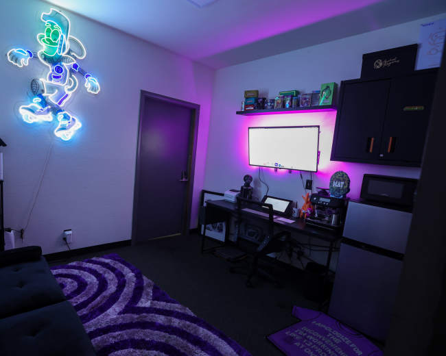 The image shows a modern office space featuring a neon light of a cartoon character, a dark couch, a desk with multiple screens, and a purple circular rug on the floor.