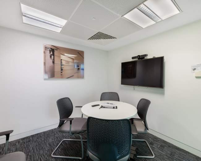 A small conference room features a round table with four chairs, a large wall-mounted screen, and a framed picture on the wall.