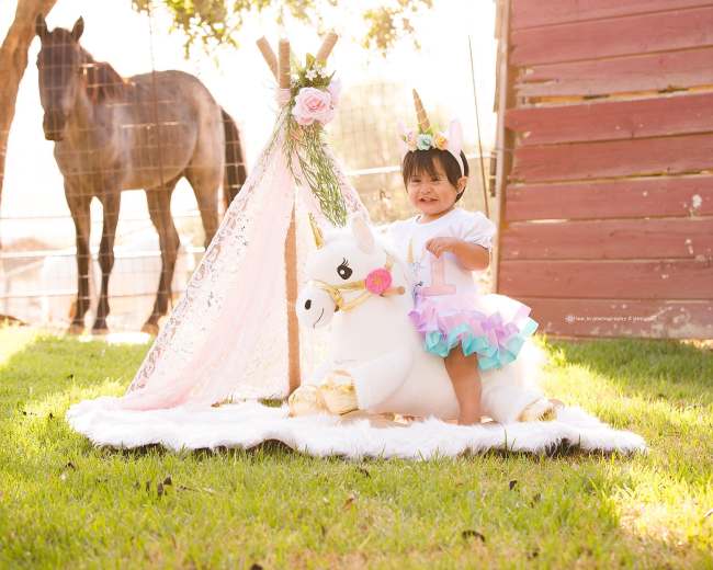 A young child stands on a rug in front of a unicorn-themed setup, with a horse in the background and sunlight filtering through the trees.