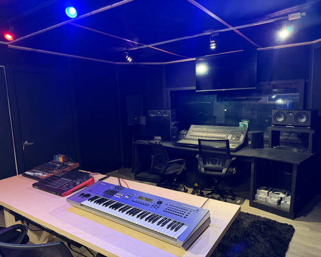 The image shows a music production studio featuring a keyboard on a desk, a mixing console at the back, and studio monitors on either side, illuminated by colored lights on the ceiling.