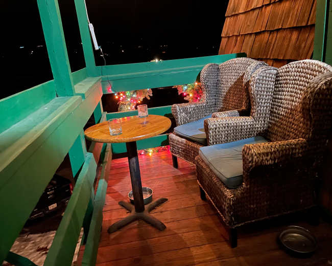 A balcony features two wicker chairs and a small wooden table, surrounded by festive lights against a nighttime backdrop.