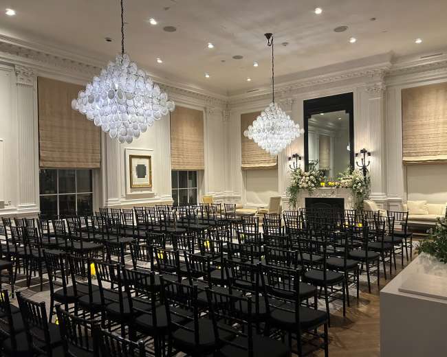 The image shows a elegantly decorated room set up with rows of black chairs facing a floral arrangement and a fireplace, illuminated by large glass chandeliers.