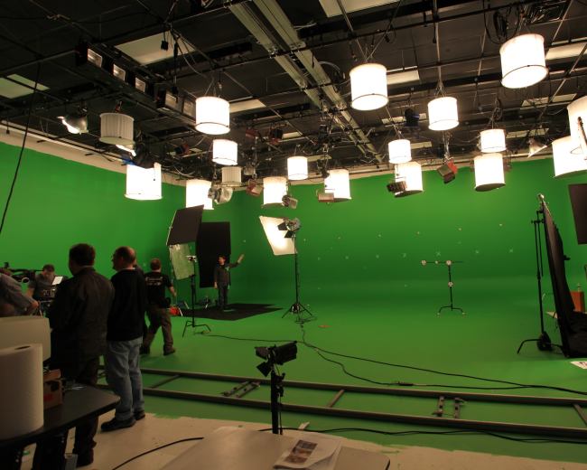 A film production set features a green screen backdrop with multiple lighting fixtures and crew members preparing for a shoot.