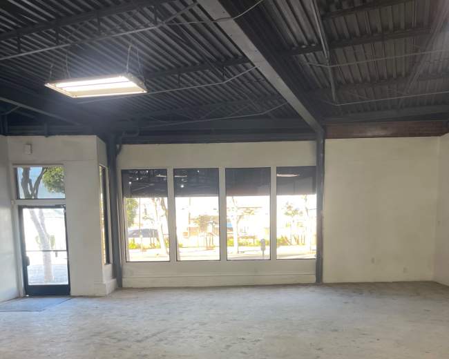 The image shows a spacious, empty commercial interior with large windows allowing natural light and a single entrance doorway.