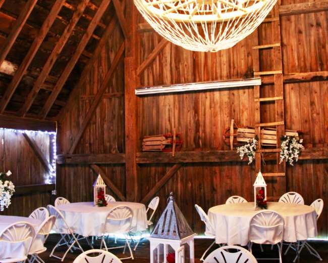 The interior of a wooden barn features round tables with white tablecloths and a large chandelier hanging from the ceiling.