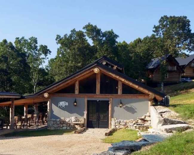 The image shows a rustic cabin with a stone and wood exterior, surrounded by trees and featuring an outdoor seating area.