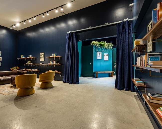 The image shows a modern, dark-colored room with a seating area featuring two yellow armchairs, a brown leather sofa, and bookshelves lined with books on the walls.